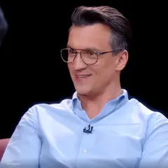 Michał Czernecki w jasnej koszuli i okularach, uśmiecha się. W tle widać fragmenty scenografii, przypominające o jego filmach, serialach i programach.