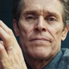 Zbliżenie na twarz Willema Dafoe, aktora znanego z wielu niezapomnianych ról w filmach. Jego dłonie są splecione przed twarzą.