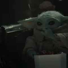 Dziecko z mandalorian film, w brązowym płaszczu, je słodycze z białego pudełka. Obok widać fragment zbroi Mandalorianina.