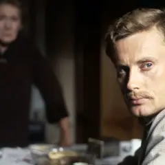 Mężczyzna z wąsami patrzy w kamerę, za nim niewyraźna postać kobiety. Scena z filmu "Panna" Wajdy.