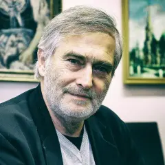 Olgierd Łukaszewicz, znany z wielu filmów, z siwą brodą i w czarnej marynarce, na tle obrazów.