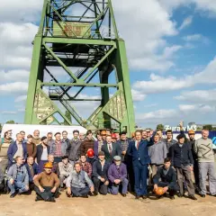 Grupa mężczyzn, w tym jeden statysta w garniturze, pozuje do zdjęcia pod zielonym dźwigiem.