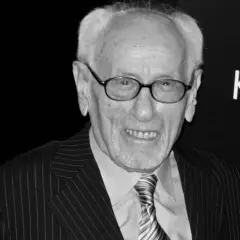 Eli Wallach, w okularach i garniturze w prążki, uśmiecha się. Ikona kina, pamiętana z niezapomnianych ról w filmach.