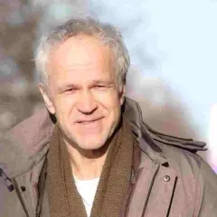 Uśmiechnięty Radosław Pazura w brązowym szaliku i kurtce, przypomina o swoich niezapomnianych rolach w polskich filmach.