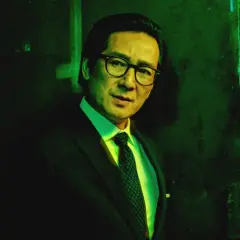 Mężczyzna w okularach i garniturze, jak z filmów ke huy quan, stoi w zielonym półmroku.