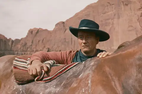 John Wayne w kapeluszu, oparty o konia, w krajobrazie przypominającym miasto bezprawia.