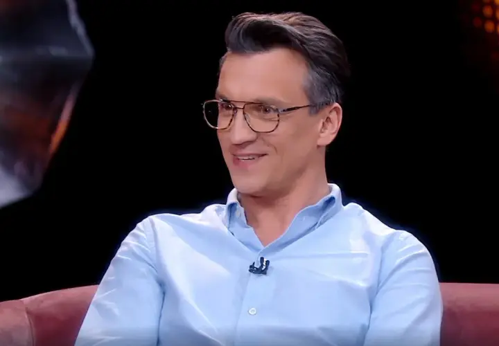 Michał Czernecki w jasnej koszuli i okularach, uśmiecha się. W tle widać fragmenty scenografii, przypominające o jego filmach, serialach i programach.