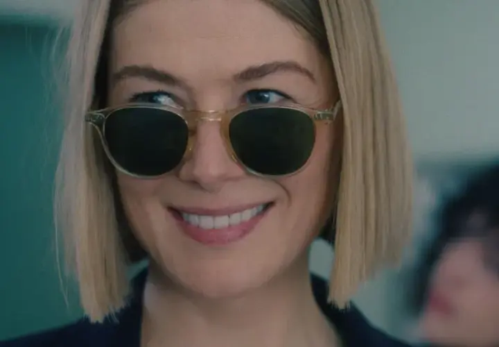 Uśmiechnięta Rosamund Pike w okularach przeciwsłonecznych, przypominająca scenę z jej filmów i seriali.