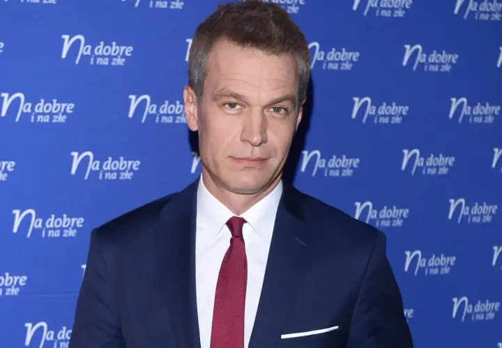 Michał Żebrowski w garniturze, znany z wielu filmów, pozuje na tle niebieskiej ścianki z napisem "Na dobre i na złe".