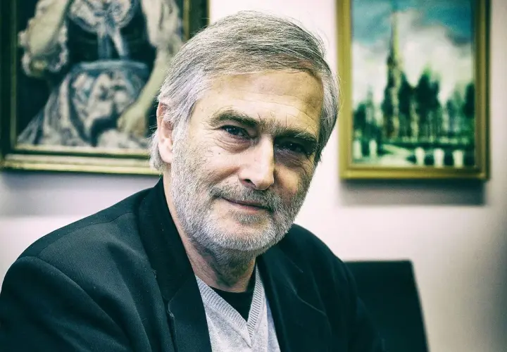 Olgierd Łukaszewicz, znany z wielu filmów, z siwą brodą i w czarnej marynarce, na tle obrazów.