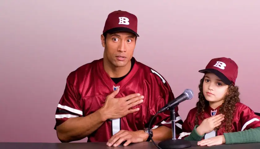 Dwayne Johnson i młoda dziewczyna w strojach drużyny sportowej, z rękami na sercu, przygotowują się do wygłoszenia przemówienia. Plan gry jest gotowy.