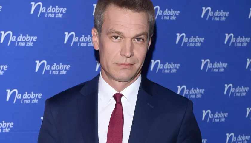 Michał Żebrowski w garniturze, znany z wielu filmów, pozuje na tle niebieskiej ścianki z napisem "Na dobre i na złe".