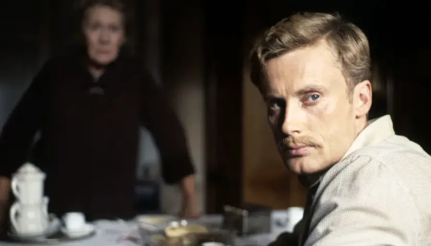 Mężczyzna z wąsami patrzy w kamerę, za nim niewyraźna postać kobiety. Scena z filmu "Panna" Wajdy.