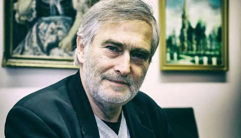 Olgierd Łukaszewicz, znany z wielu filmów, z siwą brodą i w czarnej marynarce, na tle obrazów.