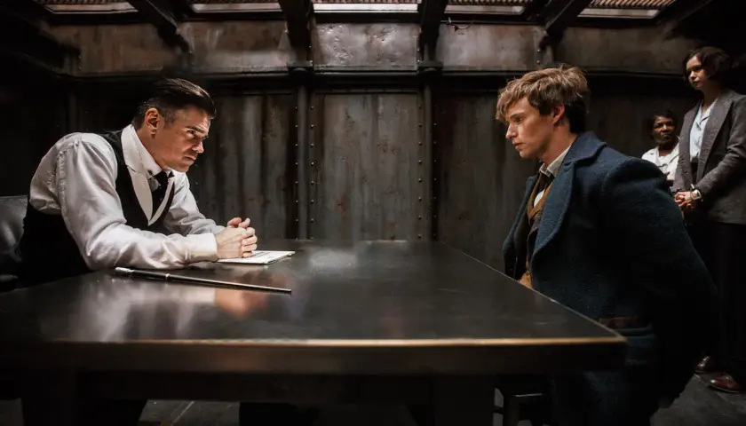 Scena z filmu "Fantastyczne zwierzęta i jak je znaleźć". Newt Scamander i Percival Graves siedzą przy stole, podczas gdy dwie kobiety stoją w tle.