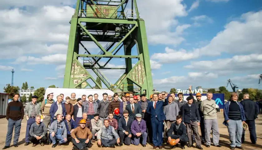 Grupa mężczyzn, w tym jeden statysta w garniturze, pozuje do zdjęcia pod zielonym dźwigiem.