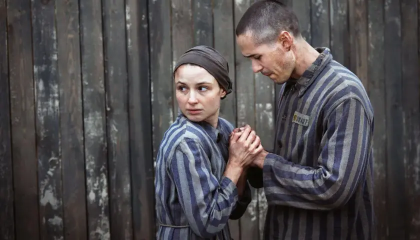 Para więźniów w pasiakach, scena z filmu o Auschwitz. Trzymają się za ręce, patrząc na siebie z nadzieją i strachem.