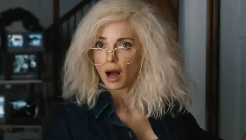 Kobieta z blond włosami i okularami, przerażona, jakby oglądała straszny film 6. W tle monitory.