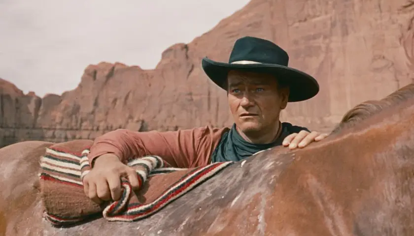 John Wayne w kapeluszu, oparty o konia, w krajobrazie przypominającym miasto bezprawia.