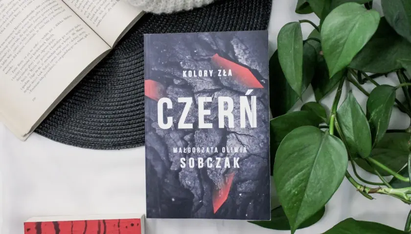 Kolory zła: Czerń. Filmowa okładka książki Małgorzaty Olwii Sobczak, otoczona zielonymi liśćmi i innymi książkami.