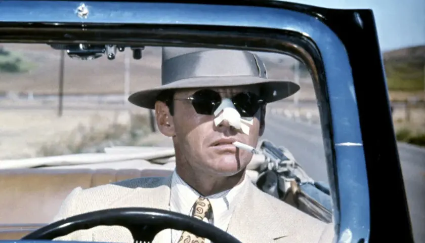 Jack Nicholson w kapeluszu i okularach przeciwsłonecznych, z plastrem na nosie, pali papierosa za kierownicą kabrioletu. Scena z filmu Polańskiego z Nicholsonem.