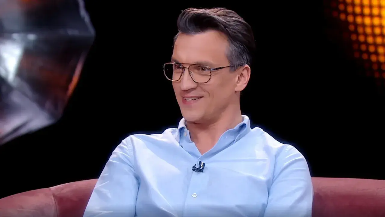 Michał Czernecki w jasnej koszuli i okularach, uśmiecha się. W tle widać fragmenty scenografii, przypominające o jego filmach, serialach i programach.