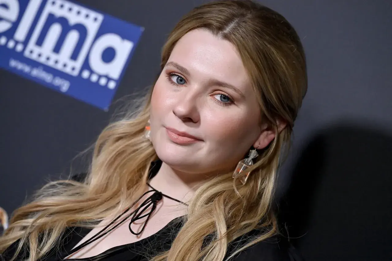 Abigail Breslin, znana z wielu filmów, z niebieskimi oczami i blond włosami, pozuje z uśmiechem.