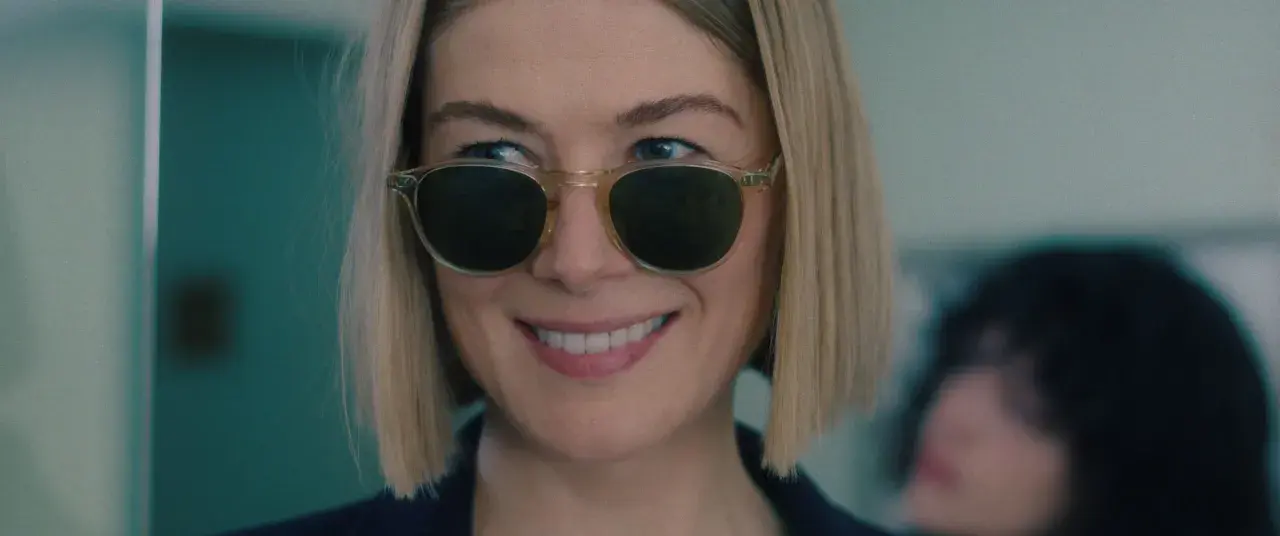 Uśmiechnięta Rosamund Pike w okularach przeciwsłonecznych, przypominająca scenę z jej filmów i seriali.