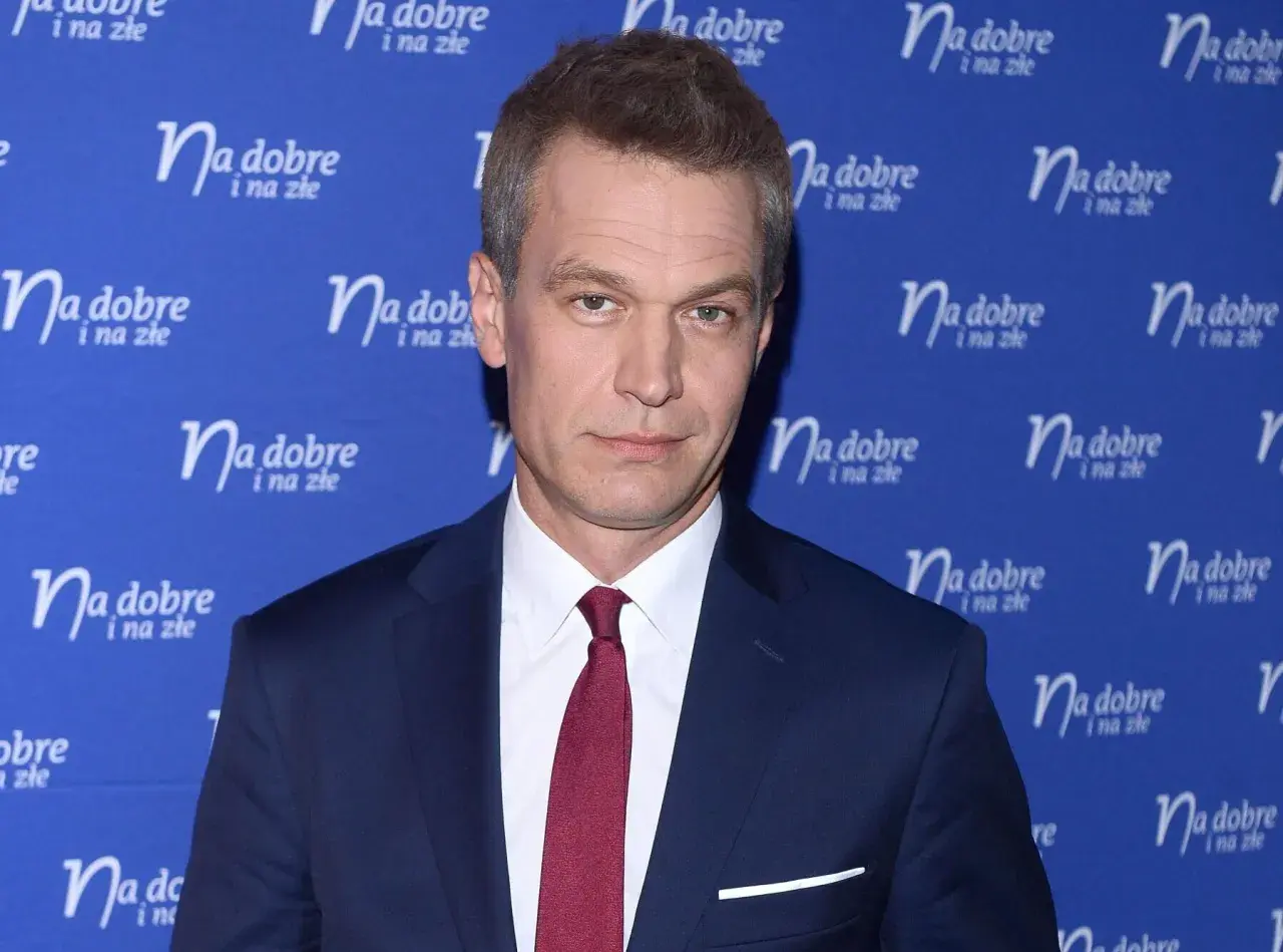 Michał Żebrowski w garniturze, znany z wielu filmów, pozuje na tle niebieskiej ścianki z napisem "Na dobre i na złe".