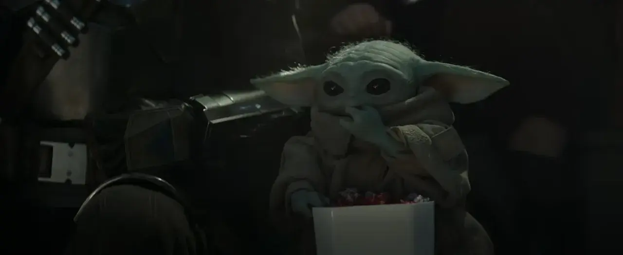 Dziecko z mandalorian film, w brązowym płaszczu, je słodycze z białego pudełka. Obok widać fragment zbroi Mandalorianina.