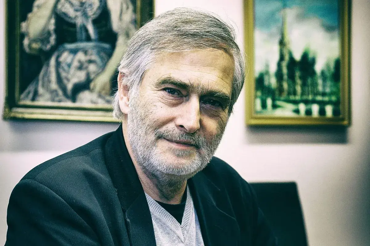 Olgierd Łukaszewicz, znany z wielu filmów, z siwą brodą i w czarnej marynarce, na tle obrazów.