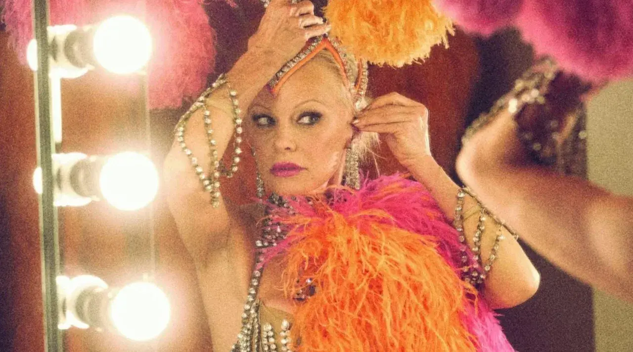 Pamela Anderson w kolorowym piórkowym stroju, przygotowująca się do występu. Jej styl przypomina kreacje z jej filmów.