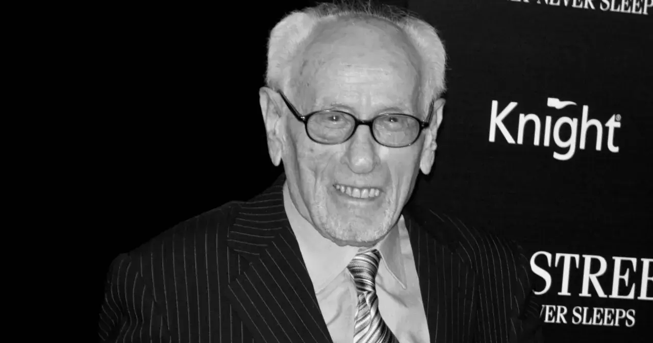 Eli Wallach, w okularach i garniturze w prążki, uśmiecha się. Ikona kina, pamiętana z niezapomnianych ról w filmach.