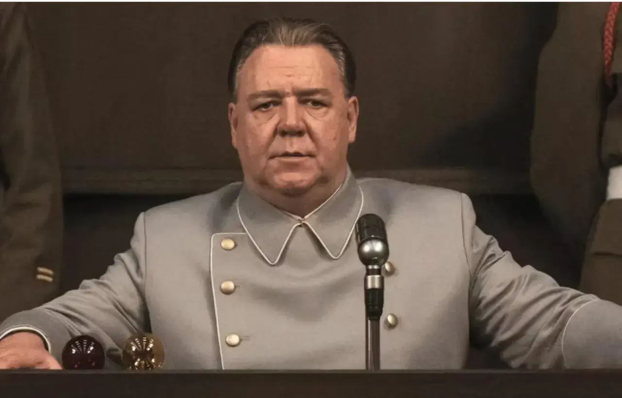 Russell Crowe w szarym mundurze, z mikrofonem przed sobą. Scena z jednego z jego filmów.