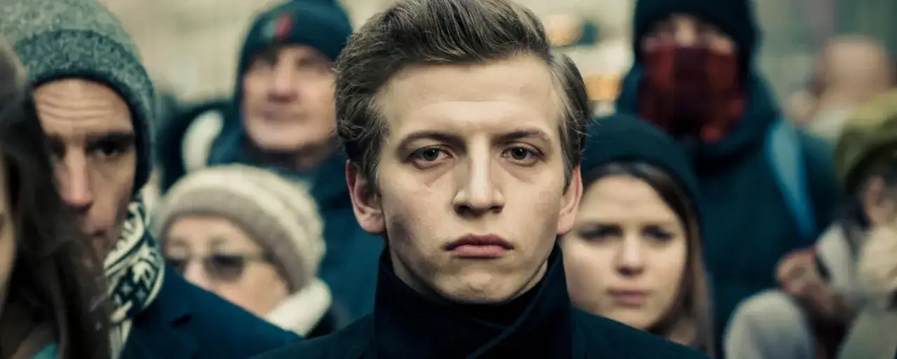 Młody mężczyzna w centrum, otoczony przez tłum. Jego wyraz twarzy sugeruje napięcie, jakby przygotowywał się do ważnej sceny w jednym z filmów z jego udziałem. Maciej Musiałowski filmy.