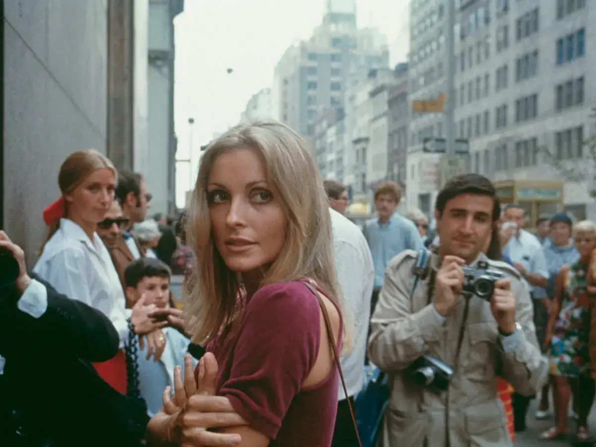 Sharon Tate, gwiazda filmów, w tłumie na ulicy. Obok niej mężczyzna z aparatem, a w tle miejski krajobraz.
