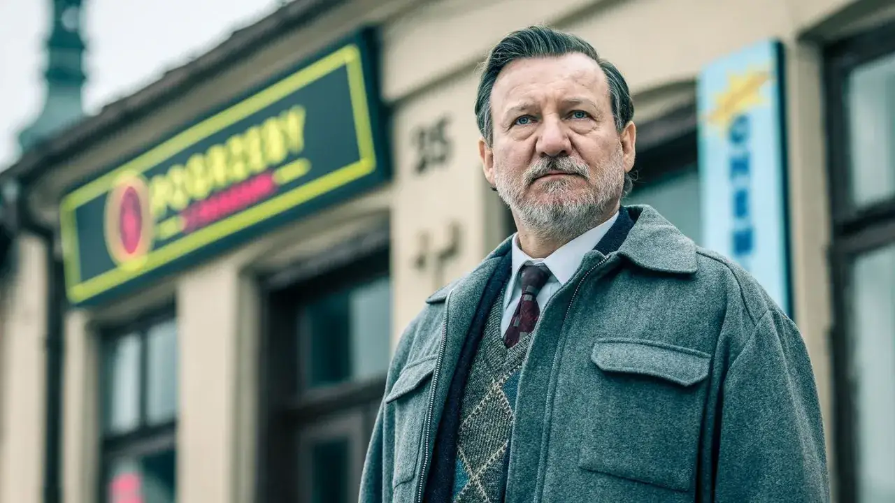 Mężczyzna w szarym płaszczu i swetrze w romby stoi przed sklepem. To scena z filmu 