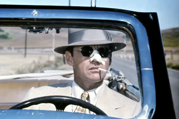 Jack Nicholson w kapeluszu i okularach przeciwsłonecznych, z plastrem na nosie, pali papierosa za kierownicą kabrioletu. Scena z filmu Polańskiego z Nicholsonem.
